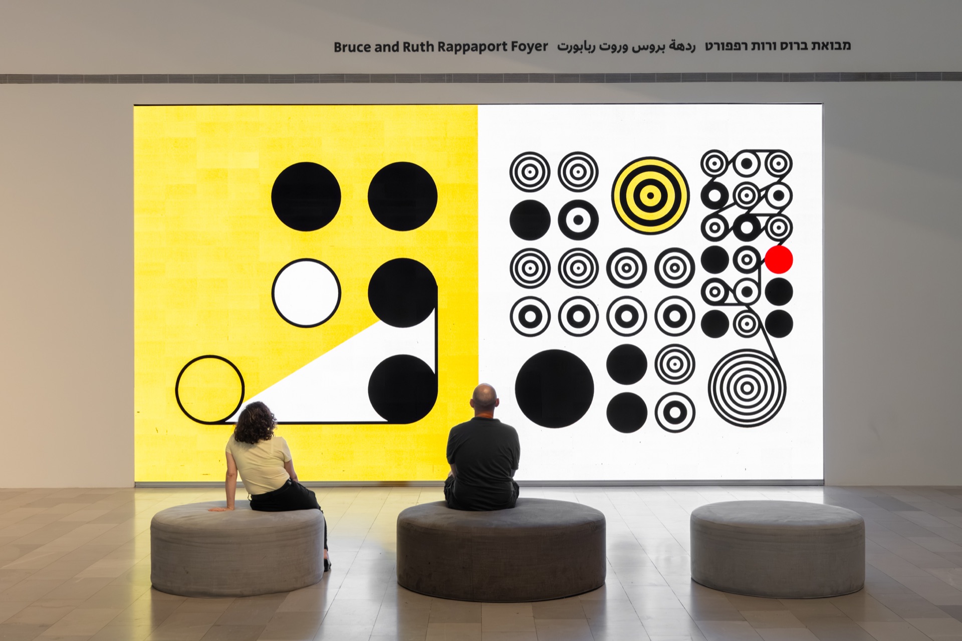 Two visitors seated before the yellow, black and white Ringers in Motion installation at the Tel Aviv Museum of Art.