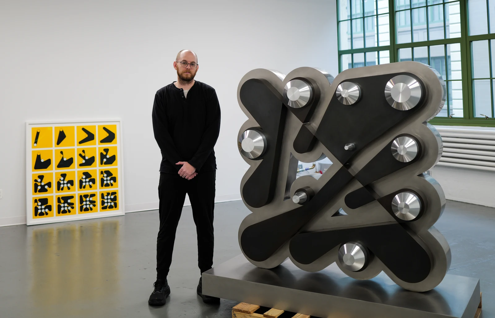 Dmitri Cherniak standing beside a large metal sculpture in the studio, with a framed grid of yellow Ringers compositions behind him.