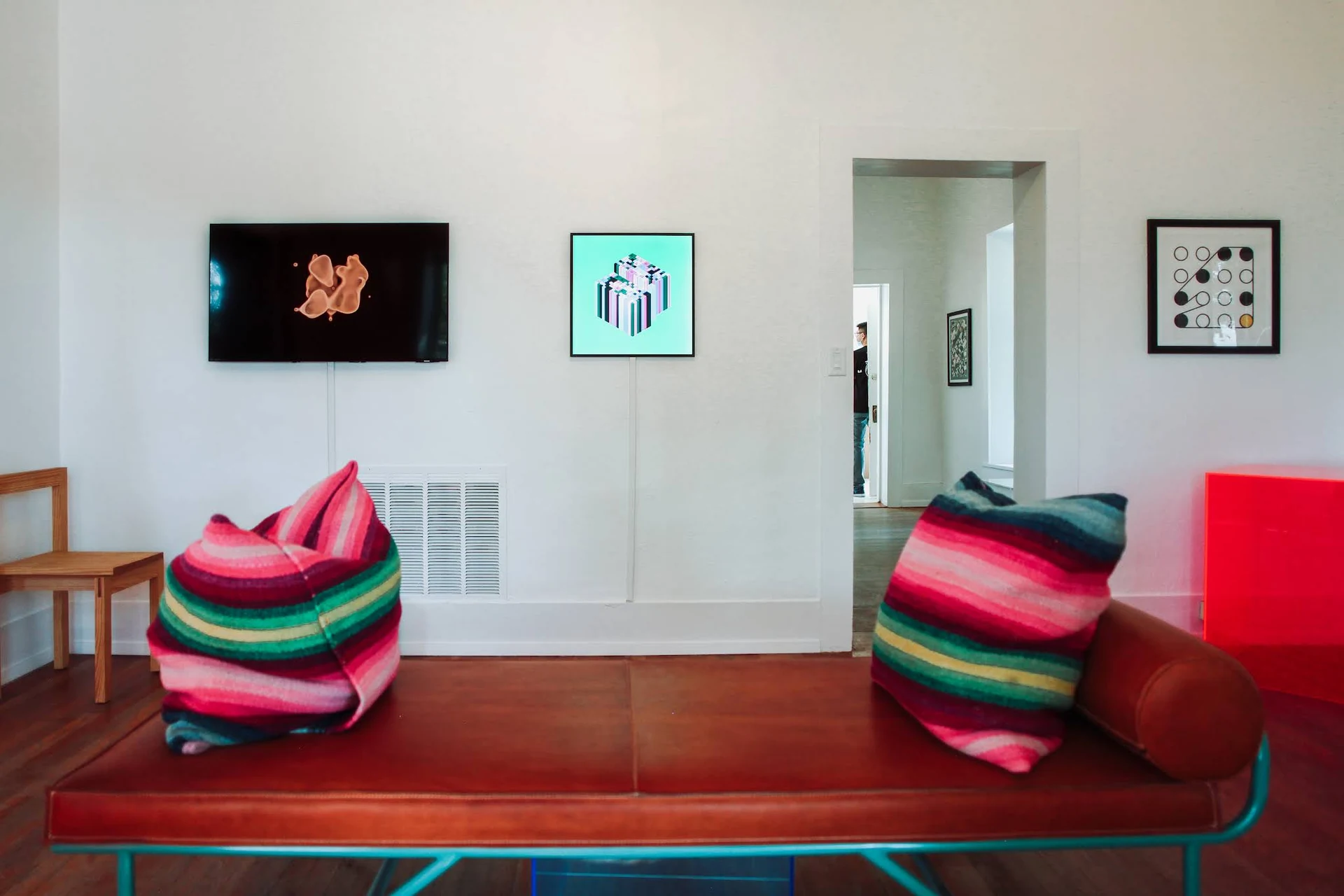 A white gallery room with a leather daybed and striped cushions in front of three works: a screen showing an orange form, a square screen with an isometric cube, and, at right beside a doorway, Ringers #0 framed in black.