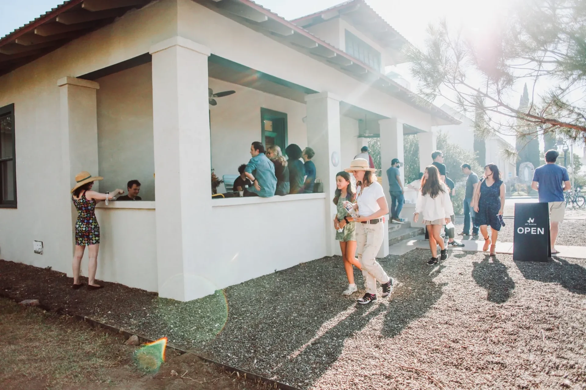 Visitors on the porch and gravel yard of the gallery, a white house with square columns, beside a black sign reading Art Blocks Open, in bright afternoon sun.