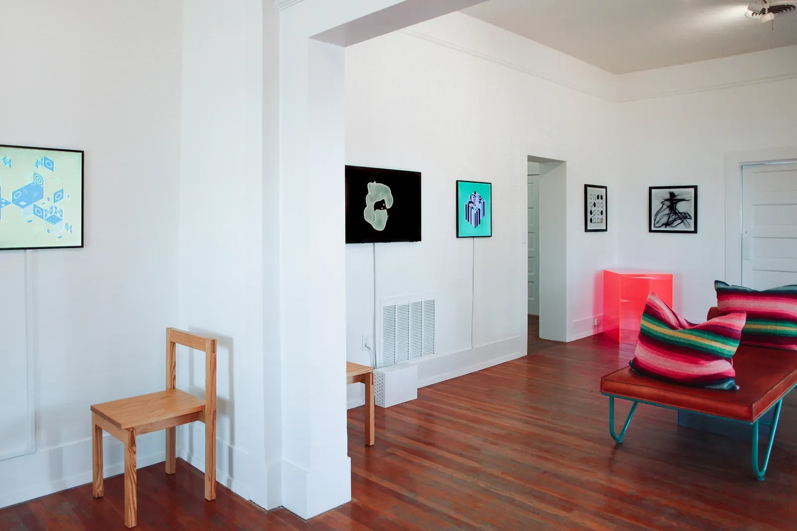 A wider view of the gallery's white rooms, with screens and framed works on the walls, a wooden chair, and a leather daybed; Ringers #0 hangs beside a doorway at the back.