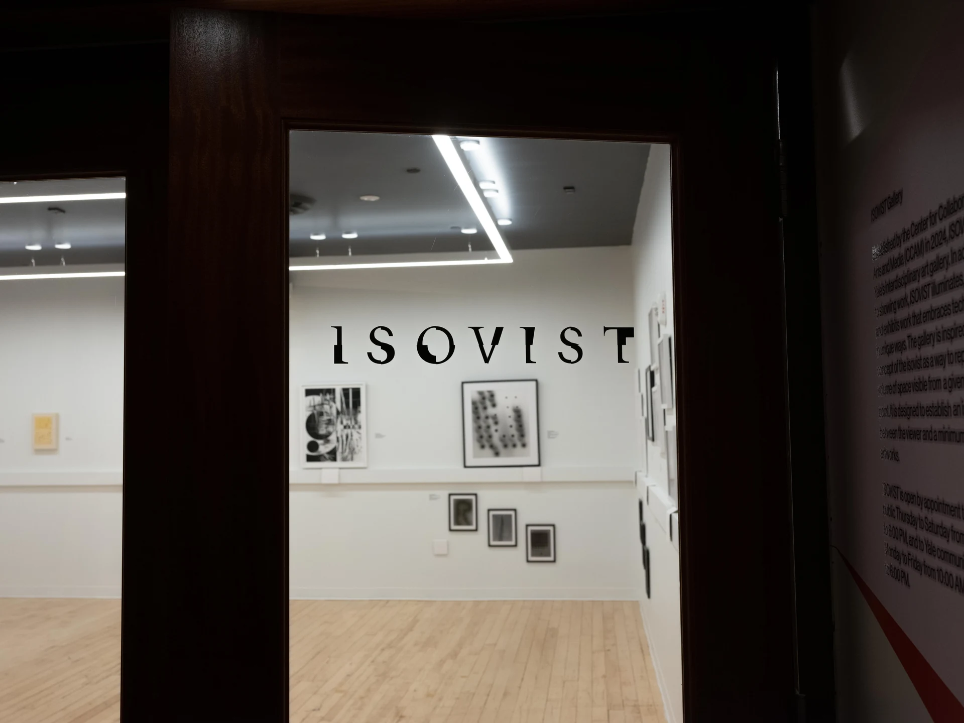 A view through a dark wooden doorway into a white gallery, where the word ISOVIST is painted in large black letters above framed black-and-white works.
