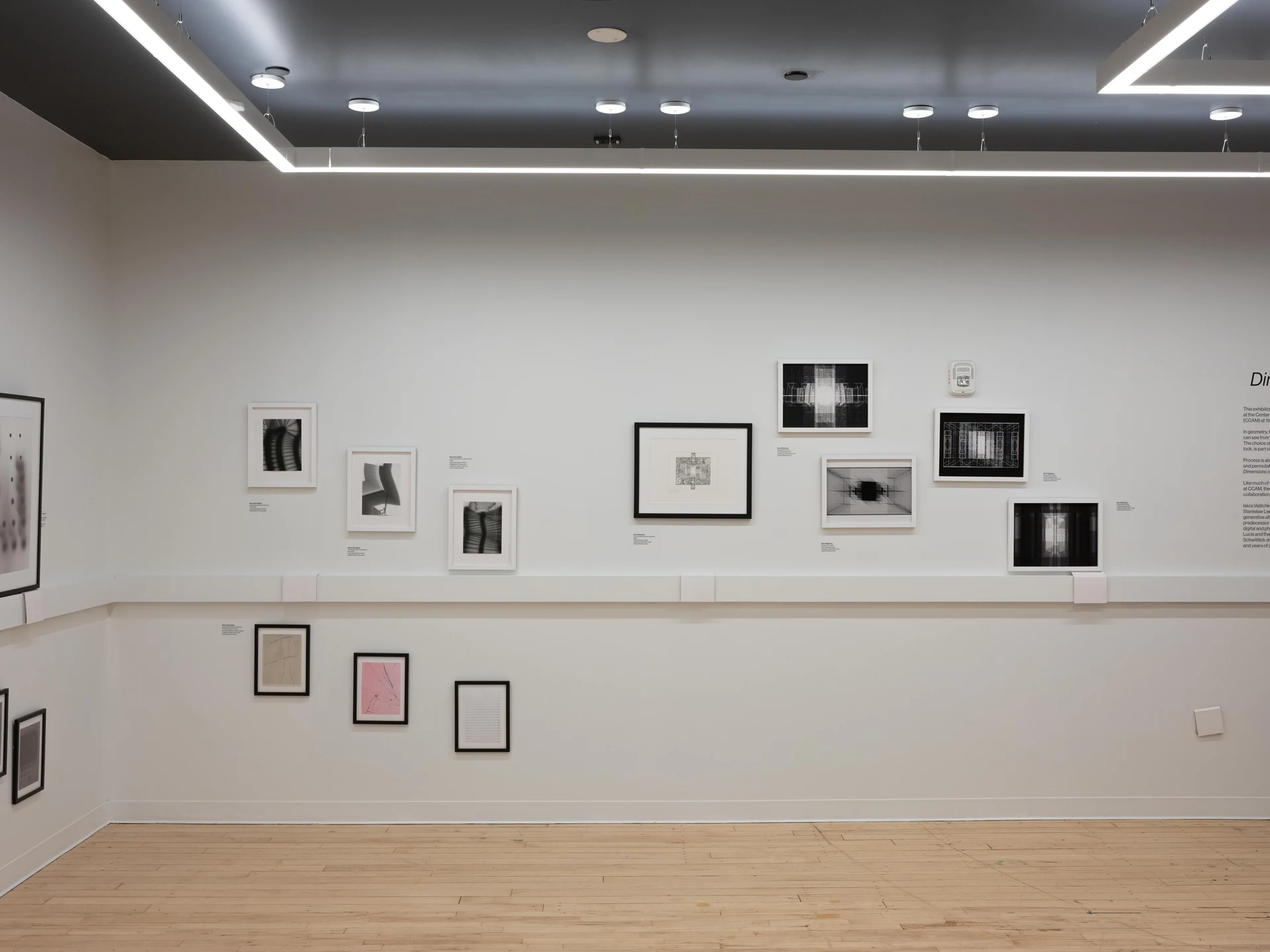 A white gallery wall hung with small framed prints and drawings in black, white, and pink, above a light wooden floor.