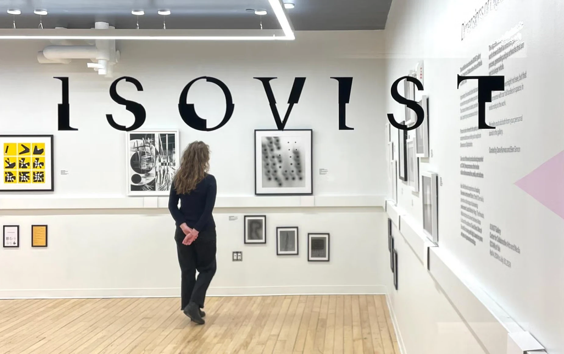 A visitor with long hair, hands clasped behind her back, stands in a white gallery beneath the word ISOVIST in large black letters. On the wall ahead hang a yellow and black grid print and a black-and-white photogram, with more framed works and the exhibition text on the wall at right.