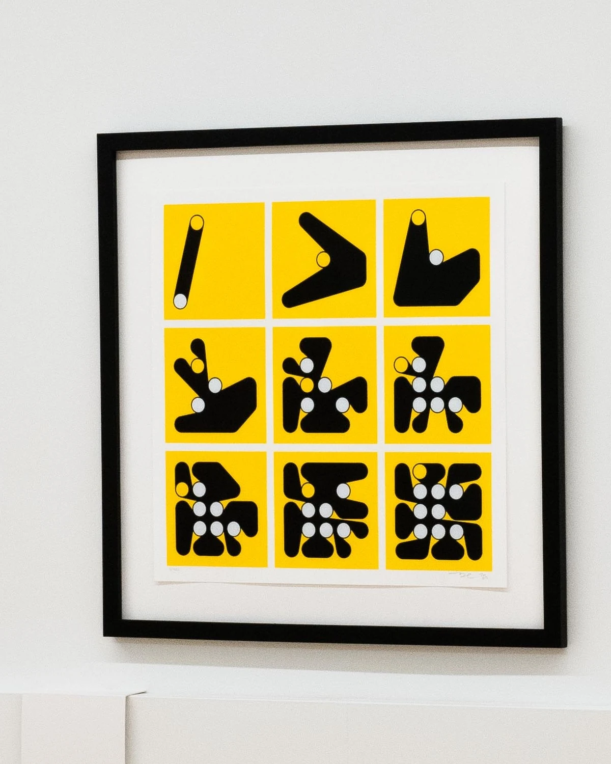 A framed print of nine yellow squares in a three-by-three grid, each with a black form wrapped around white pegs, growing from a single bar at top left to a dense cluster at bottom right.