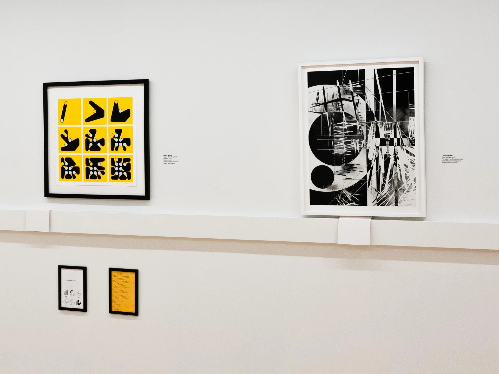 A white gallery wall with two framed prints above a ledge: at left, a yellow and black three-by-three grid in a black frame; at right, a black-and-white photogram of circles, lines, and white strokes in a white frame, each with a wall label. Below the ledge hang two small framed pages, one with diagrams of peg grids and one of code on yellow paper.