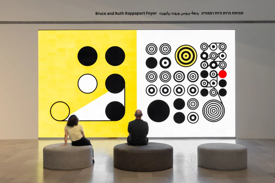 Two people seated before large yellow, black, and white circular compositions on the installation screen.