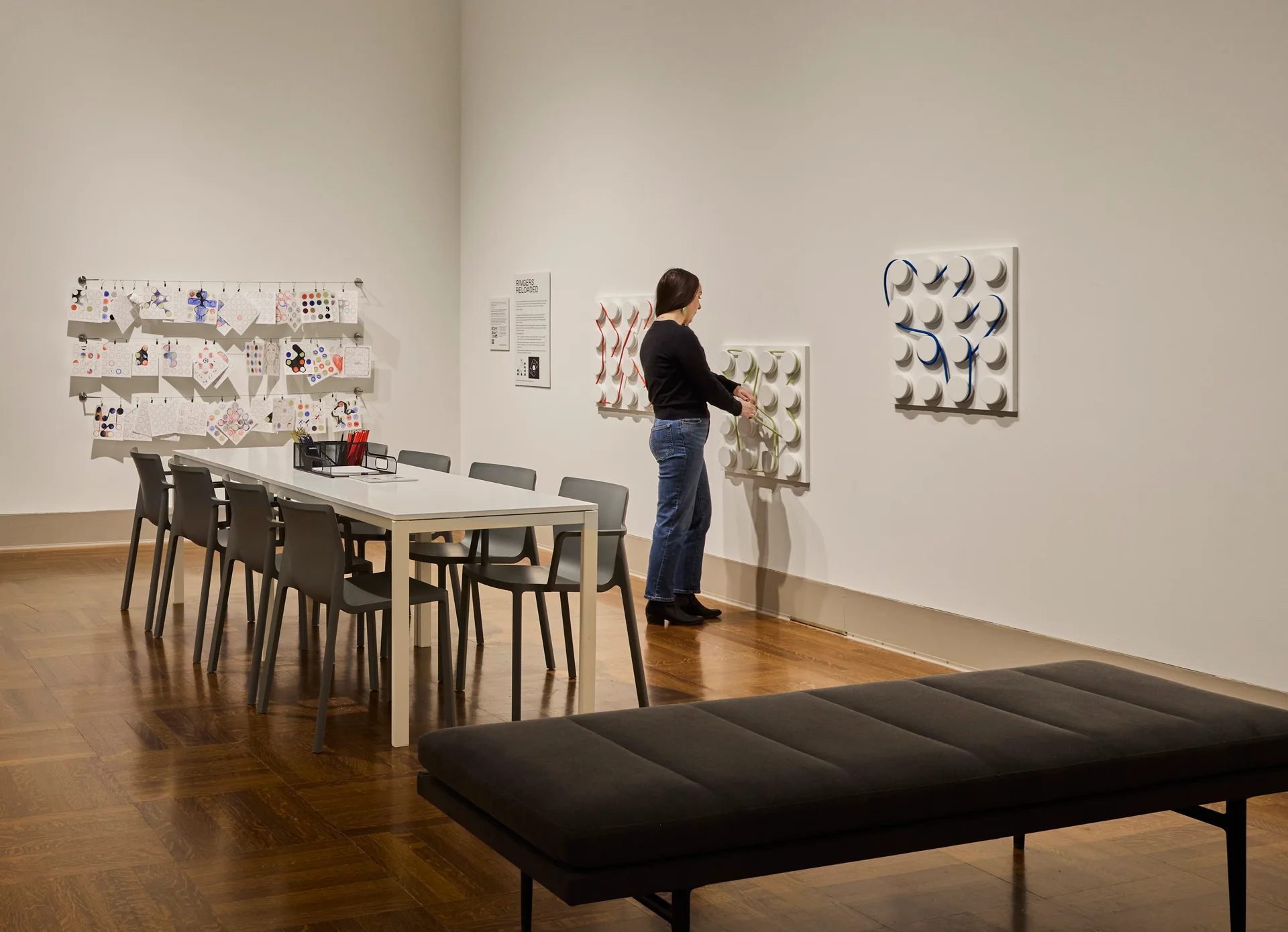 A visitor wraps string around large wall-mounted pegs beside a drawing table and a display of visitor drawings.