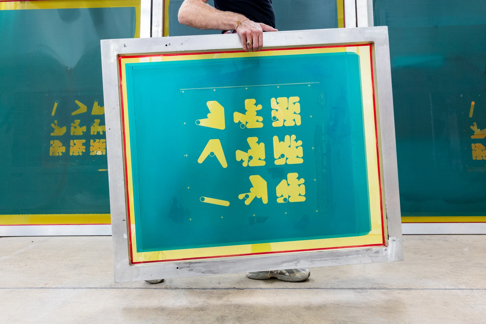 A person holds a screenprinting frame with yellow Ringers forms against green mesh, with more prepared screens behind it.