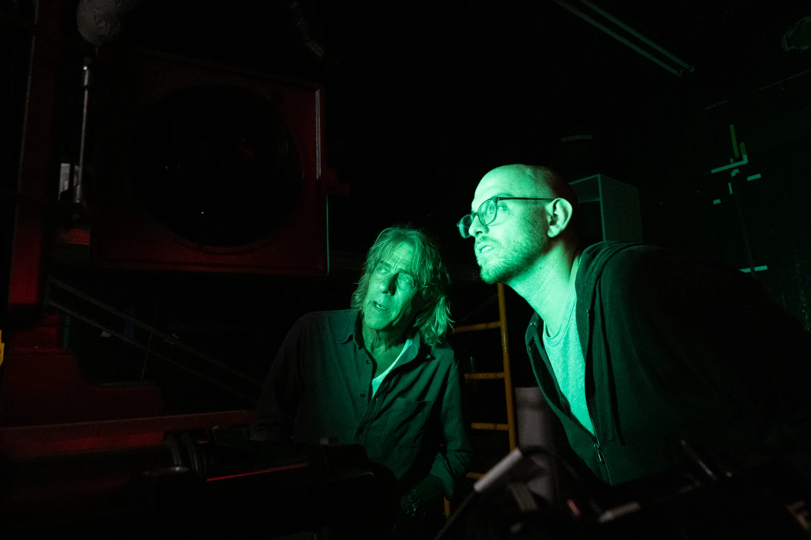 Two people examining photographic equipment under green darkroom light.