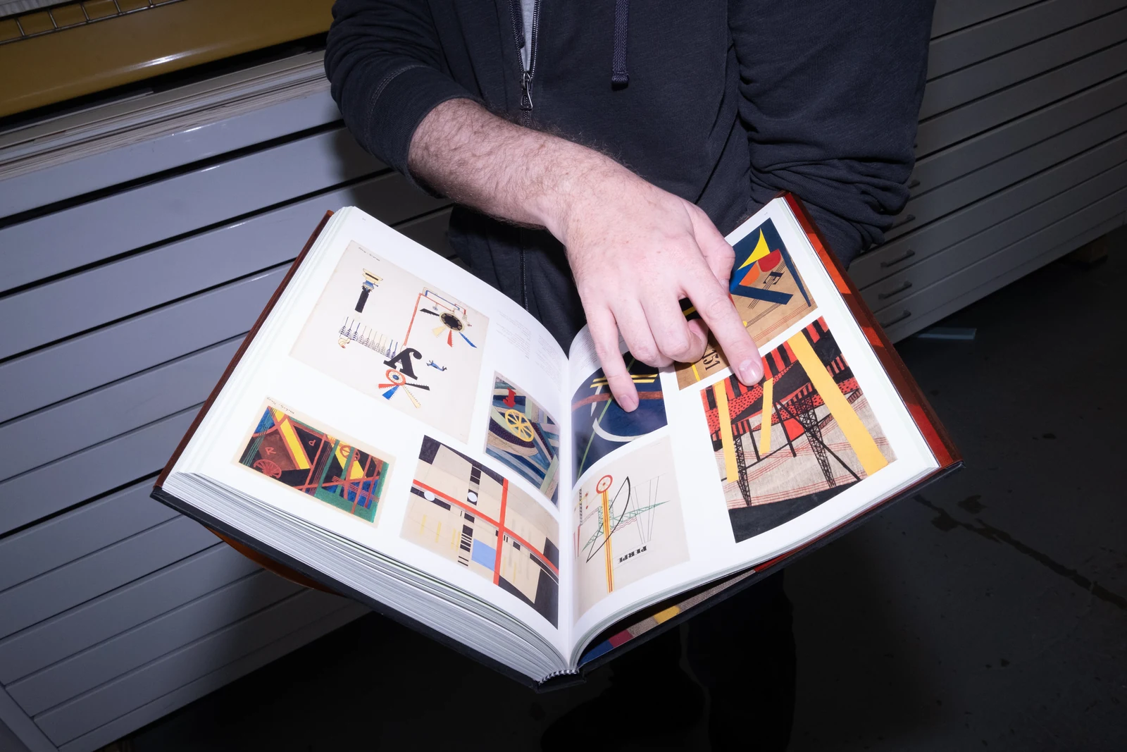 An open art book with reproductions of geometric compositions, held above studio drawers.