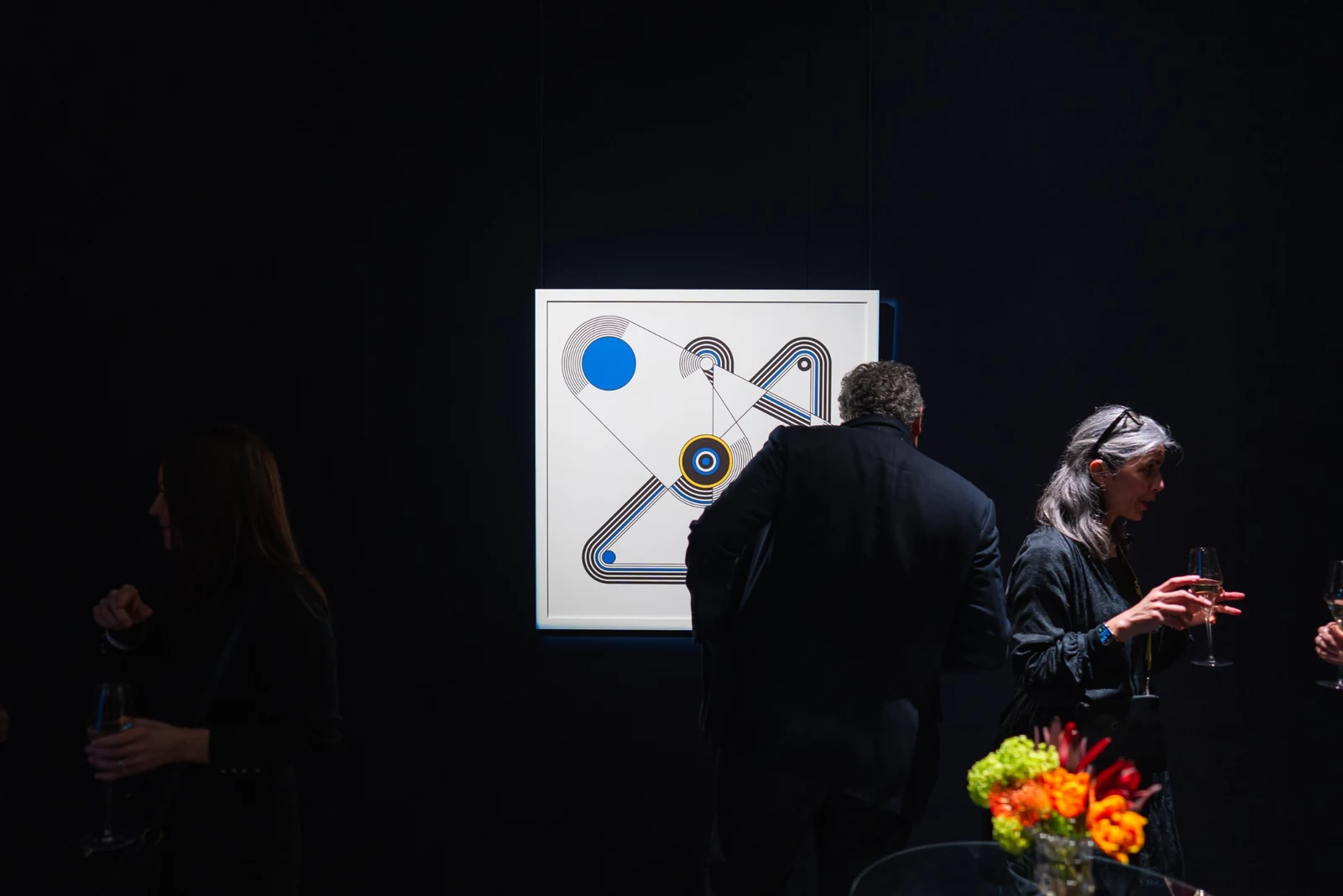 The framed print hangs alone on a dark wall while visitors talk nearby, with flowers in the foreground.