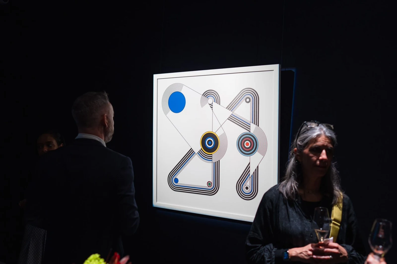 Visitors holding glasses stand in a dark room in front of the lit, framed print.